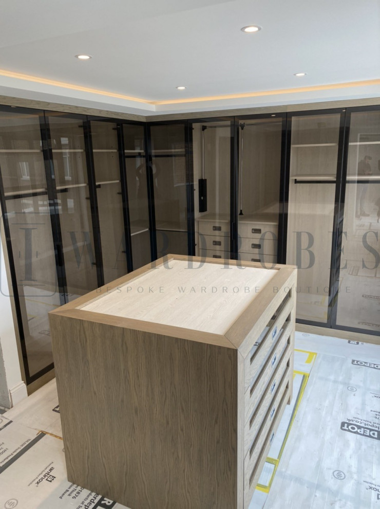 This is an example of a large contemporary built-in wardrobe in London with glass-front cabinets and light wood cabinets.