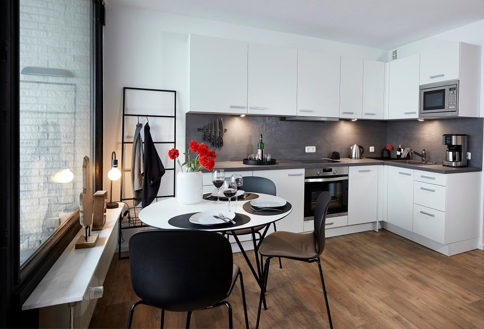 Mittelgroße Moderne Wohnküche ohne Insel in L-Form mit Einbauwaschbecken, flächenbündigen Schrankfronten, weißen Schränken, Küchenrückwand in Grau, Küchengeräten aus Edelstahl, braunem Holzboden, braunem Boden und grauer Arbeitsplatte in Hamburg