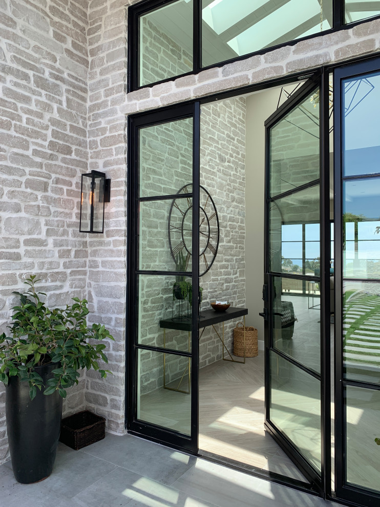 Photo of a mid-sized beach style front door in Orange County with beige walls, porcelain floors, a pivot front door, a black front door, beige floor and brick walls.