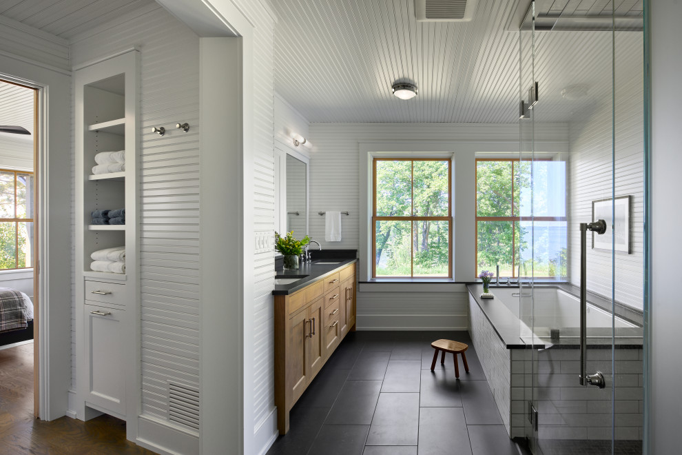 Example of a beach style master white tile black floor, double-sink and wall paneling corner shower design in Minneapolis with recessed-panel cabinets, medium tone wood cabinets, an undermount tub, white walls, an undermount sink, a hinged shower door, black countertops and a built-in vanity
