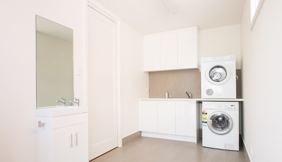 Small contemporary u-shaped dedicated laundry room in Melbourne with an undermount sink, flat-panel cabinets, white cabinets, limestone benchtops, white walls, ceramic floors and a stacked washer and dryer.