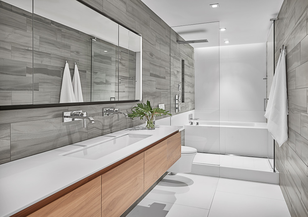 This is an example of a mid-sized contemporary master wet room bathroom in Chicago with flat-panel cabinets, medium wood cabinets, a wall-mount toilet, limestone, an undermount sink, solid surface benchtops, white floor, a hinged shower door, white benchtops, an undermount tub and gray tile.