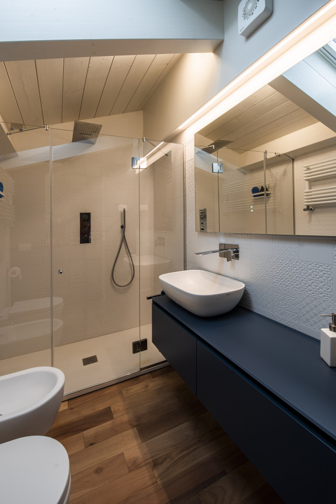 Пример оригинального дизайна: маленькая ванная комната с плоскими фасадами, синими фасадами, раздельным унитазом, белой плиткой, керамогранитной плиткой, белыми стенами, деревянным полом, настольной раковиной, стеклянной столешницей, коричневым полом, душем с распашными дверями, синей столешницей, тумбой под одну раковину, подвесной тумбой и деревянным потолком для на участке и в саду