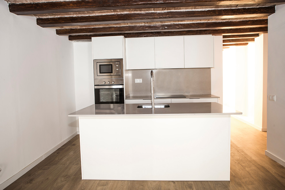 Mid-sized country galley open plan kitchen in Barcelona with a single-bowl sink, white cabinets, grey splashback, stainless steel appliances, medium hardwood floors, with island and brown floor.
