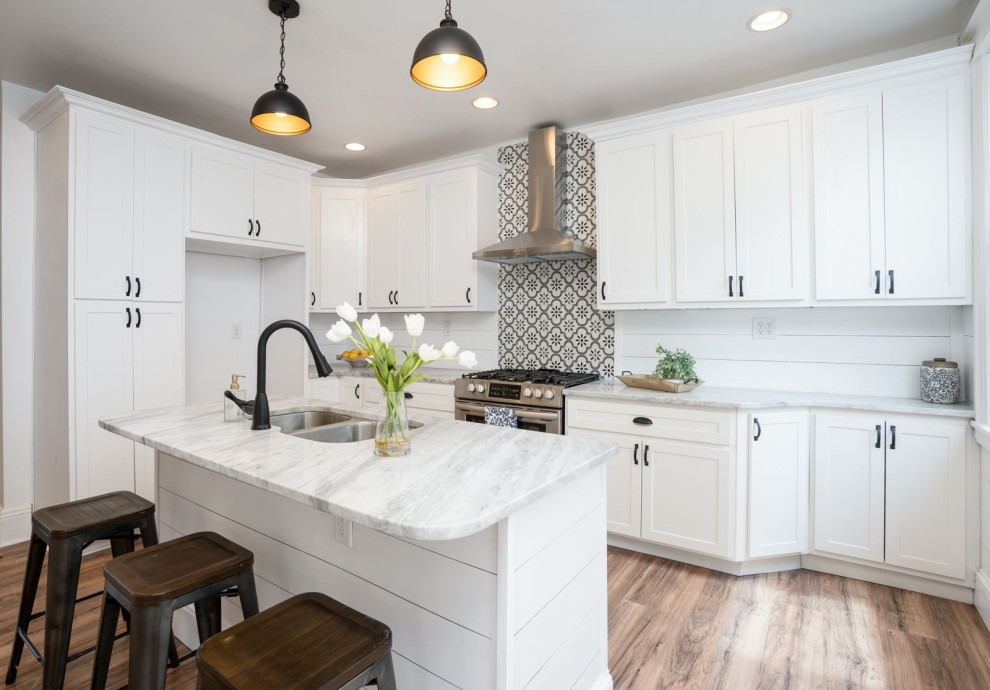 This is an example of a mid-sized country galley eat-in kitchen in St Louis with a drop-in sink, shaker cabinets, white cabinets, marble benchtops, white splashback, shiplap splashback, stainless steel appliances, light hardwood floors, with island, brown floor, grey benchtop and wood.