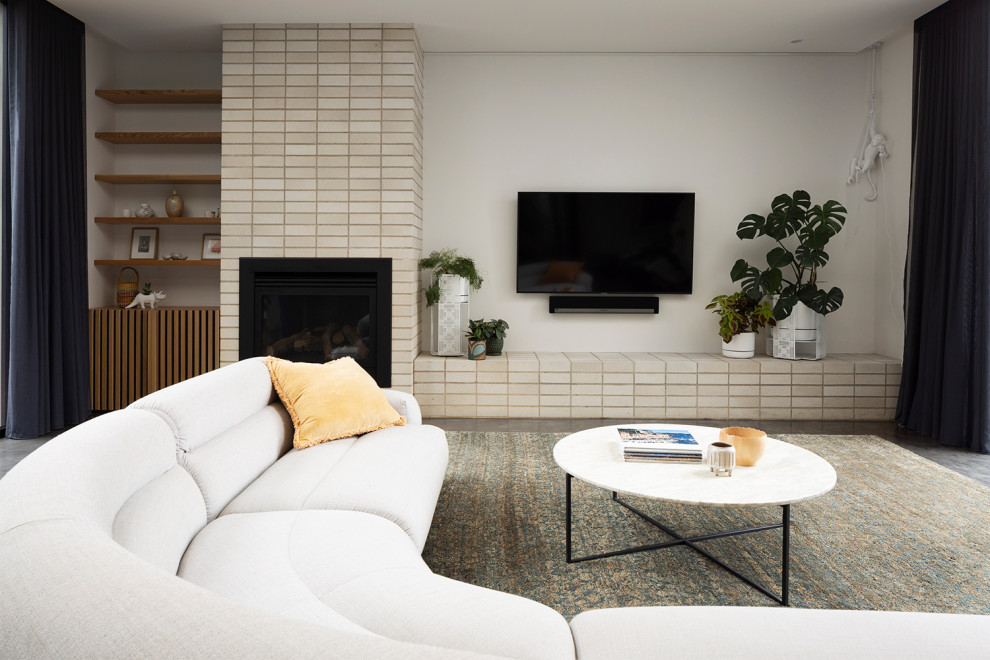 Cette image montre un grand salon design ouvert avec un mur blanc, sol en béton ciré, une cheminée standard, un manteau de cheminée en brique, un téléviseur fixé au mur et un sol gris.