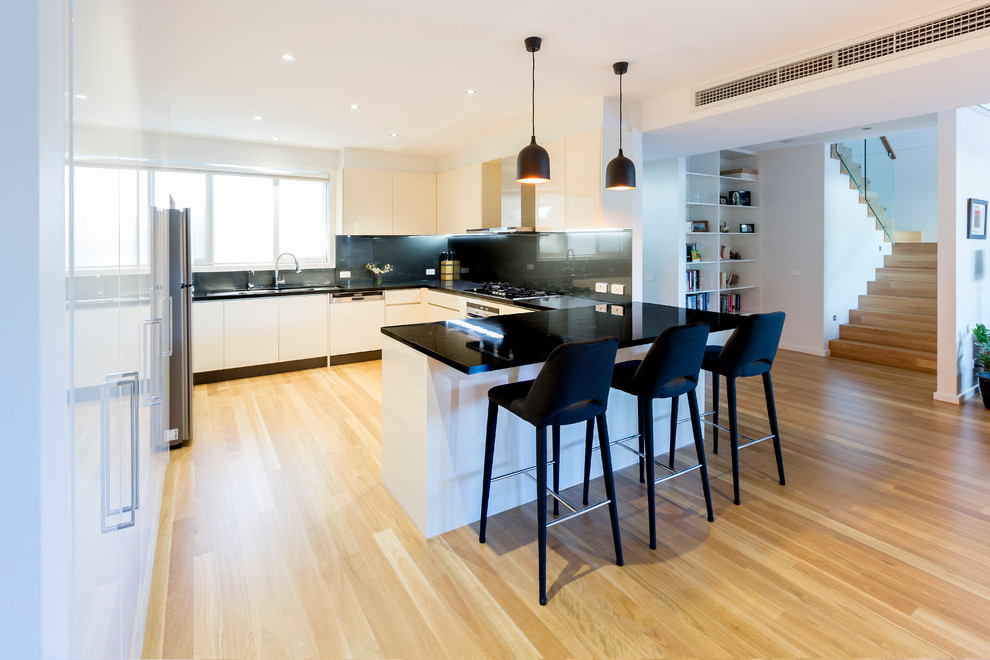 Large contemporary u-shaped kitchen in Geelong with a double-bowl sink, flat-panel cabinets, beige cabinets, quartz benchtops, metallic splashback, glass sheet splashback, black appliances, light hardwood floors and no island.