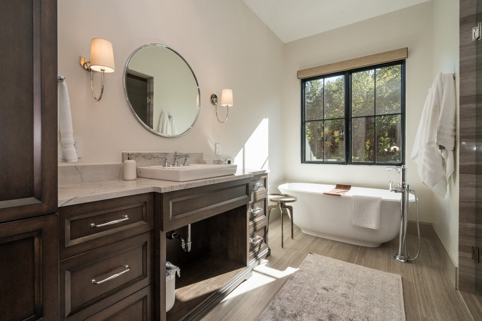 This is an example of a transitional master bathroom in St Louis with recessed-panel cabinets, dark wood cabinets, a freestanding tub, an alcove shower, white walls, wood-look tile, a vessel sink, marble benchtops, brown floor, a hinged shower door, multi-coloured benchtops, a single vanity and a built-in vanity.