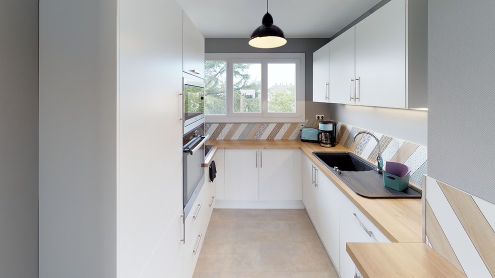 This is an example of a mid-sized modern u-shaped separate kitchen in Angers with an undermount sink, beaded inset cabinets, white cabinets, wood benchtops, multi-coloured splashback, timber splashback, black appliances, vinyl floors, grey floor and beige benchtop.