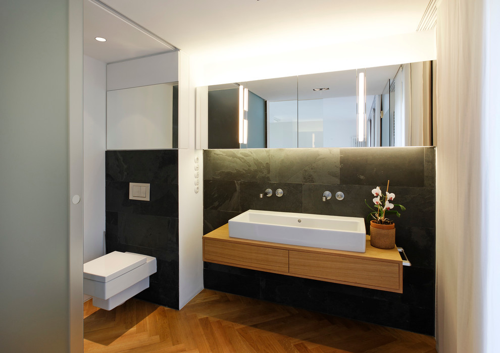 Badezimmer With flächenbündigen Schrankfronten, Wandtoilette, grauen Fliesen, schwarzen Fliesen, weißer Wandfarbe, Aufsatzwaschbecken, Waschtisch aus Holz, hellbraunen Holzschränken, braunem Holzboden, Einbaubadewanne, bodengleicher Dusche, Steinfliesen, beigem Boden and Falttür-Duschabtrennung in Hamburg