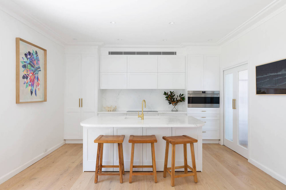 Mid-sized contemporary kitchen pantry in Sydney with a drop-in sink, recessed-panel cabinets, white cabinets, solid surface benchtops, white splashback, stone slab splashback, stainless steel appliances, light hardwood floors, with island, beige floor and white benchtop.