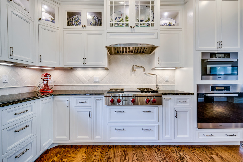 Mid-sized transitional l-shaped medium tone wood floor and brown floor open concept kitchen photo in Boston with a farmhouse sink, recessed-panel cabinets, white cabinets, granite countertops, beige backsplash, ceramic backsplash, stainless steel appliances and an island