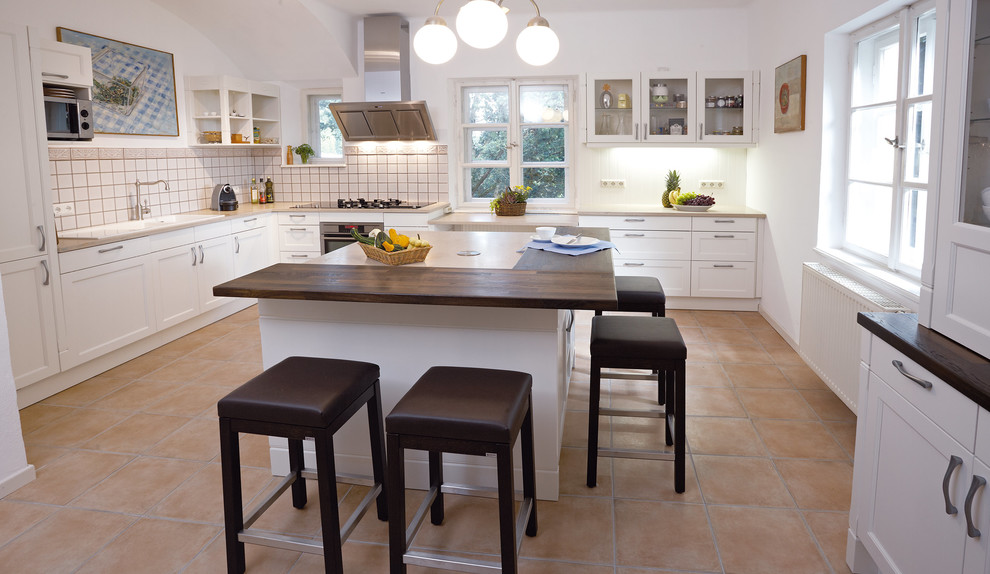 Design ideas for a mid-sized country l-shaped eat-in kitchen in San Francisco with glass-front cabinets, white cabinets, laminate benchtops, ceramic splashback, white appliances, with island, a drop-in sink, white splashback, terra-cotta floors and beige floor.