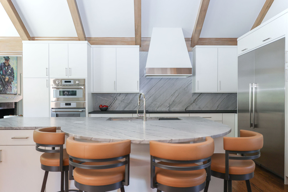 Example of a mid-sized transitional u-shaped medium tone wood floor, brown floor and vaulted ceiling open concept kitchen design with an undermount sink, flat-panel cabinets, white cabinets, solid surface countertops, gray backsplash, marble backsplash, stainless steel appliances, an island and black countertops