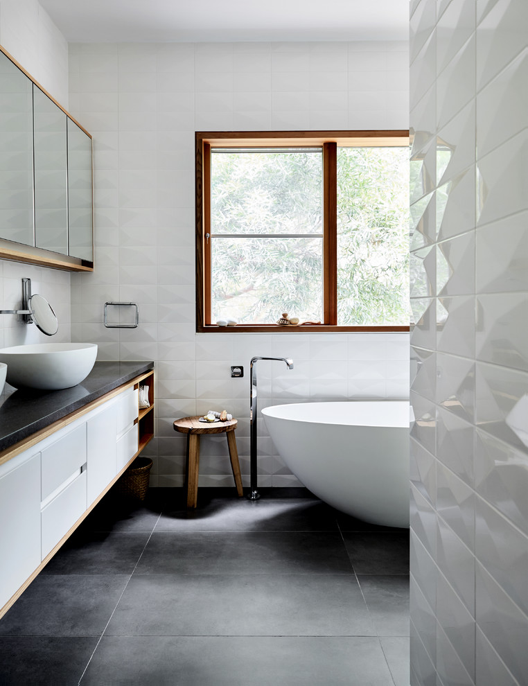 Inspiration for a mid-sized contemporary master bathroom in Melbourne with white cabinets, a freestanding tub, white tile, ceramic tile, white walls, a vessel sink, black floor, flat-panel cabinets and black benchtops.