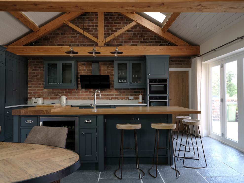 Mid-sized country l-shaped open plan kitchen in Cheshire with shaker cabinets, brick splashback, stainless steel appliances, with island and black floor.