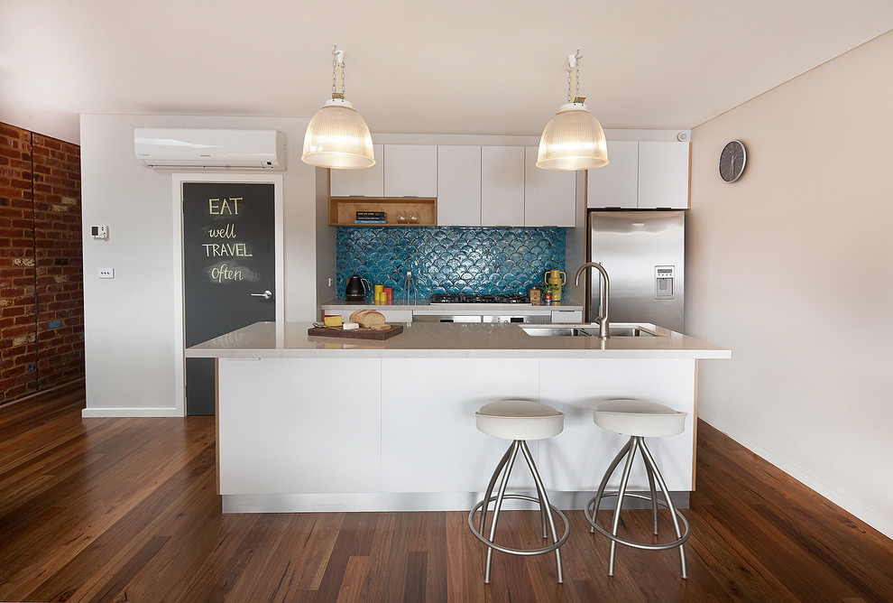 This is an example of a mid-sized contemporary single-wall eat-in kitchen in Melbourne with a double-bowl sink, flat-panel cabinets, white cabinets, blue splashback, ceramic splashback, stainless steel appliances, medium hardwood floors, with island, quartz benchtops, brown floor and white benchtop.