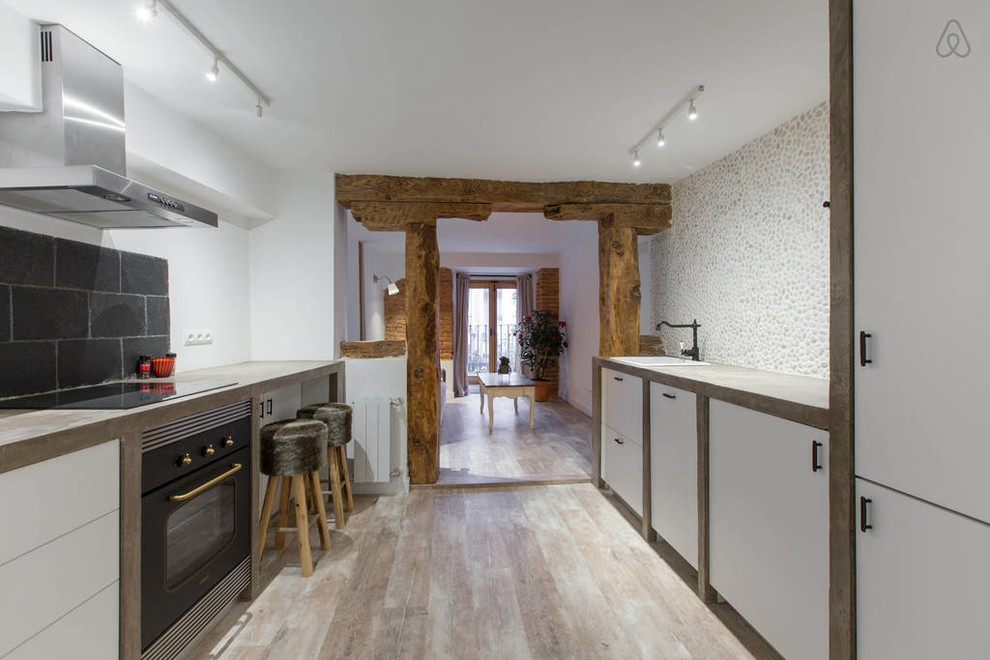 Mid-sized transitional galley open plan kitchen in Madrid with a drop-in sink, black appliances, no island, flat-panel cabinets, white cabinets, concrete benchtops, black splashback, stone tile splashback and light hardwood floors.