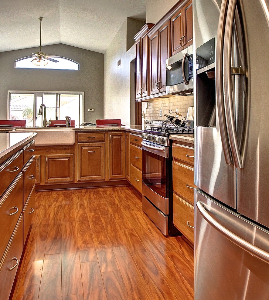 Inspiration for a mid-sized l-shaped medium tone wood floor eat-in kitchen remodel in Phoenix with a farmhouse sink, raised-panel cabinets, medium tone wood cabinets, beige backsplash, stone tile backsplash, stainless steel appliances and an island