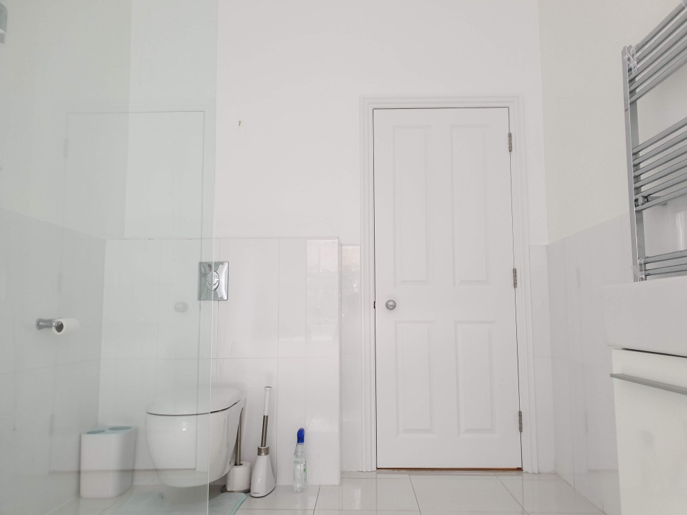 Photo of a large modern kids bathroom in London with flat-panel cabinets, white cabinets, a freestanding tub, an open shower, a wall-mount toilet, white tile, porcelain tile, white walls, pebble tile floors, a drop-in sink, marble benchtops, white floor, an open shower, white benchtops, a single vanity, a built-in vanity and recessed.