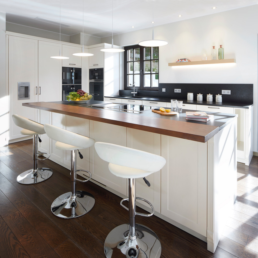 Photo of a transitional l-shaped kitchen in Barcelona with shaker cabinets, white cabinets, black splashback, black appliances, dark hardwood floors, with island, brown floor, a single-bowl sink and black benchtop.