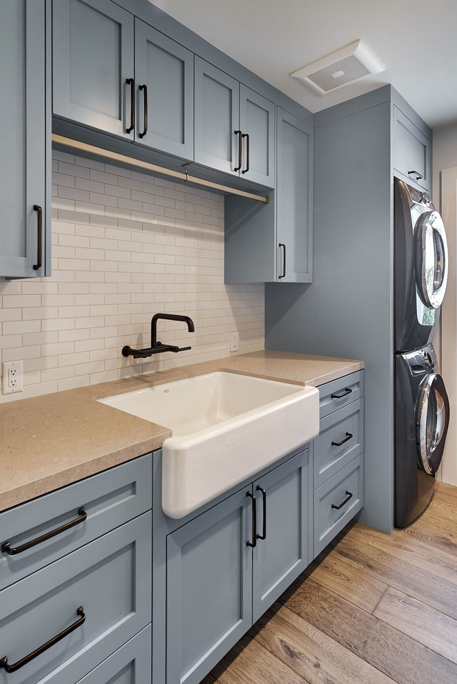 Photo of a mid-sized traditional galley dedicated laundry room in San Francisco with a farmhouse sink, shaker cabinets, blue cabinets, quartz benchtops, beige walls, medium hardwood floors, a stacked washer and dryer, brown floor and beige benchtop.