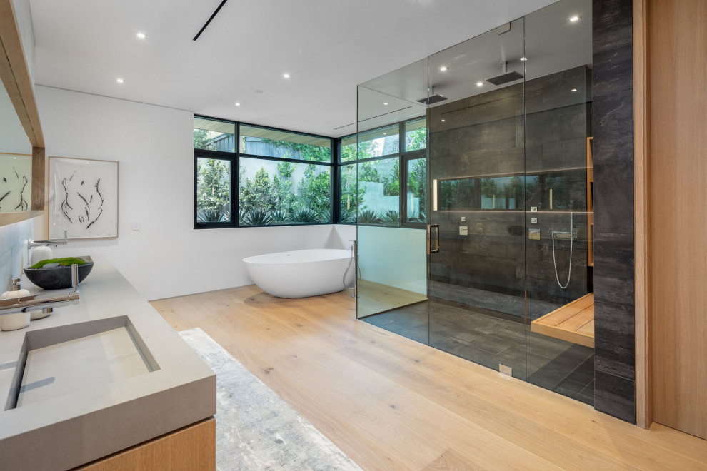 This is an example of a contemporary bathroom in Orange County with a freestanding tub, a corner shower, gray tile, white walls, light hardwood floors, an integrated sink, beige floor, a hinged shower door, grey benchtops and a double vanity.