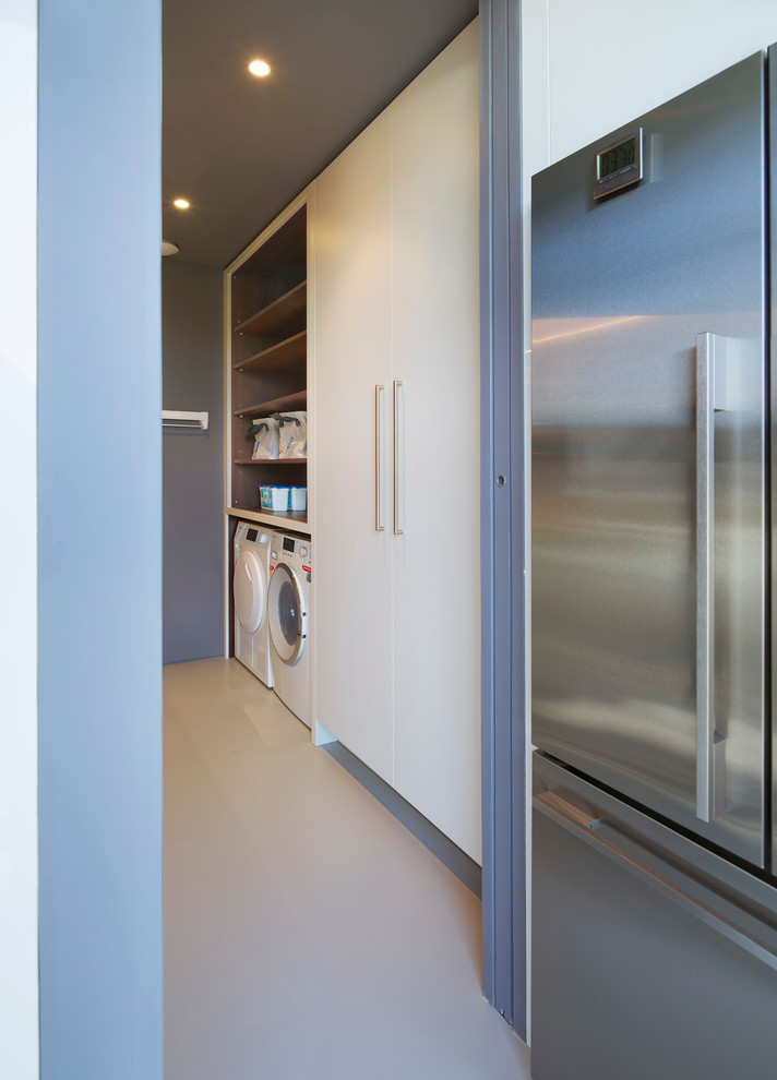 This is an example of a mid-sized contemporary galley utility room in London with a double-bowl sink, flat-panel cabinets, white cabinets, solid surface benchtops, blue walls, a side-by-side washer and dryer, beige floor and white benchtop.