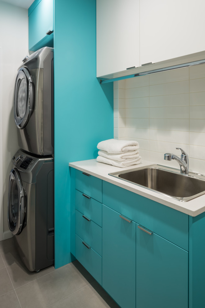 Photo of a mid-sized midcentury galley dedicated laundry room in Vancouver with a drop-in sink, flat-panel cabinets, turquoise cabinets, quartzite benchtops, white walls, porcelain floors, a stacked washer and dryer, grey floor and white benchtop.