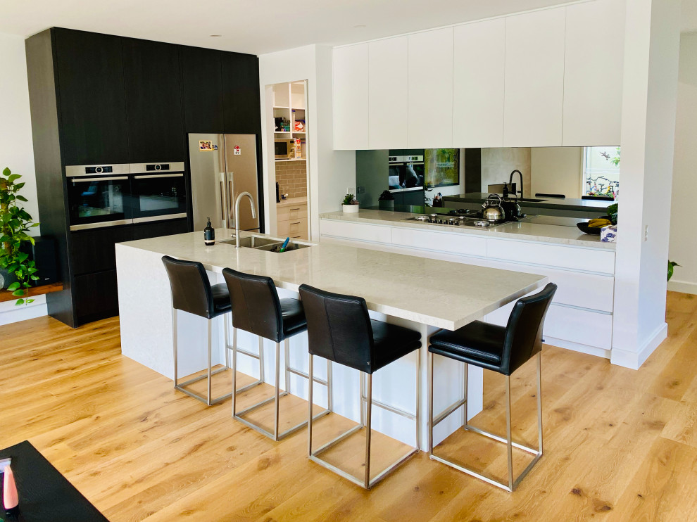 Photo of a mid-sized modern galley eat-in kitchen in Melbourne with an integrated sink, flat-panel cabinets, white cabinets, quartz benchtops, metallic splashback, mirror splashback, stainless steel appliances, light hardwood floors, with island, yellow floor and beige benchtop.