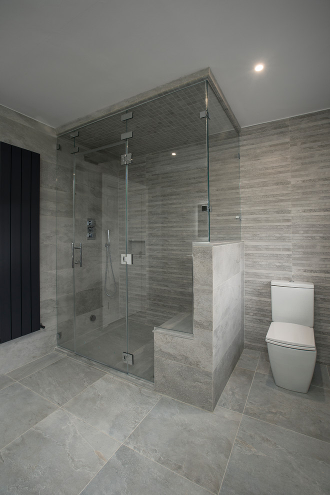 This is an example of a large contemporary master bathroom in Dublin with a curbless shower, a one-piece toilet, gray tile, grey walls, a wall-mount sink, grey floor, a hinged shower door and grey benchtops.