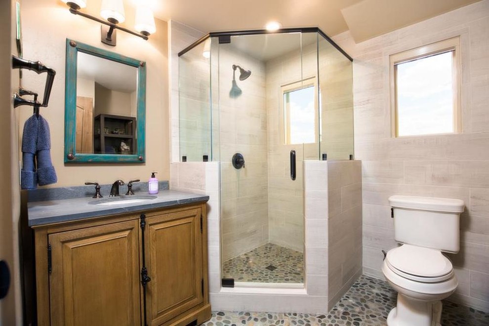 This is an example of a mid-sized country 3/4 bathroom in San Francisco with furniture-like cabinets, light wood cabinets, a corner shower, a one-piece toilet, beige tile, porcelain tile, beige walls, pebble tile floors, a drop-in sink, soapstone benchtops, grey floor, a hinged shower door and grey benchtops.