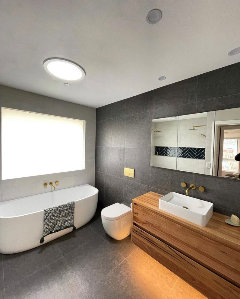 Photo of a midcentury master bathroom in Orange County with a freestanding tub, gray tile, stone tile, grey walls, slate floors, wood benchtops, grey floor, brown benchtops, a single vanity and a floating vanity.