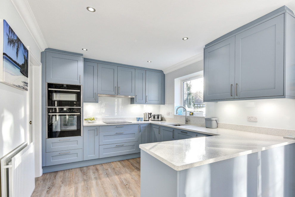 Inspiration for a mid-sized modern u-shaped eat-in kitchen in Sussex with an undermount sink, shaker cabinets, blue cabinets, quartzite benchtops, white splashback, glass sheet splashback, black appliances, light hardwood floors, no island, brown floor and white benchtop.