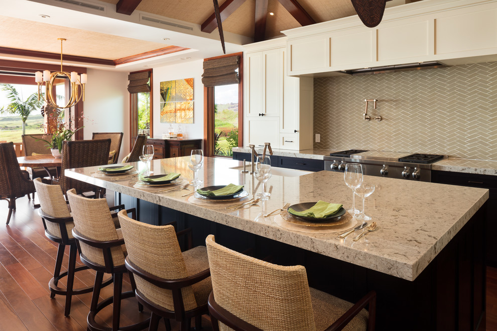 This is an example of a mid-sized tropical u-shaped eat-in kitchen in Hawaii with a farmhouse sink, shaker cabinets, white cabinets, quartz benchtops, beige splashback, glass tile splashback, stainless steel appliances, medium hardwood floors, with island, brown floor and grey benchtop.