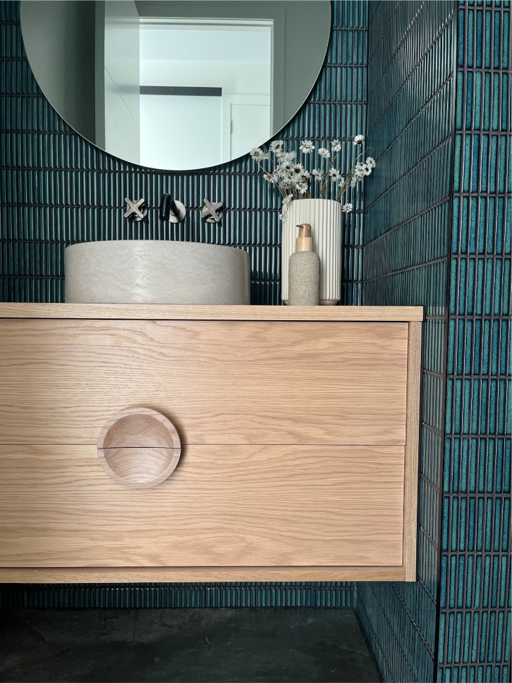 This is an example of a mid-sized contemporary powder room in Hamilton with recessed-panel cabinets, light wood cabinets, a one-piece toilet, green tile, matchstick tile, white walls, concrete floors, a vessel sink, wood benchtops, black floor and a floating vanity.