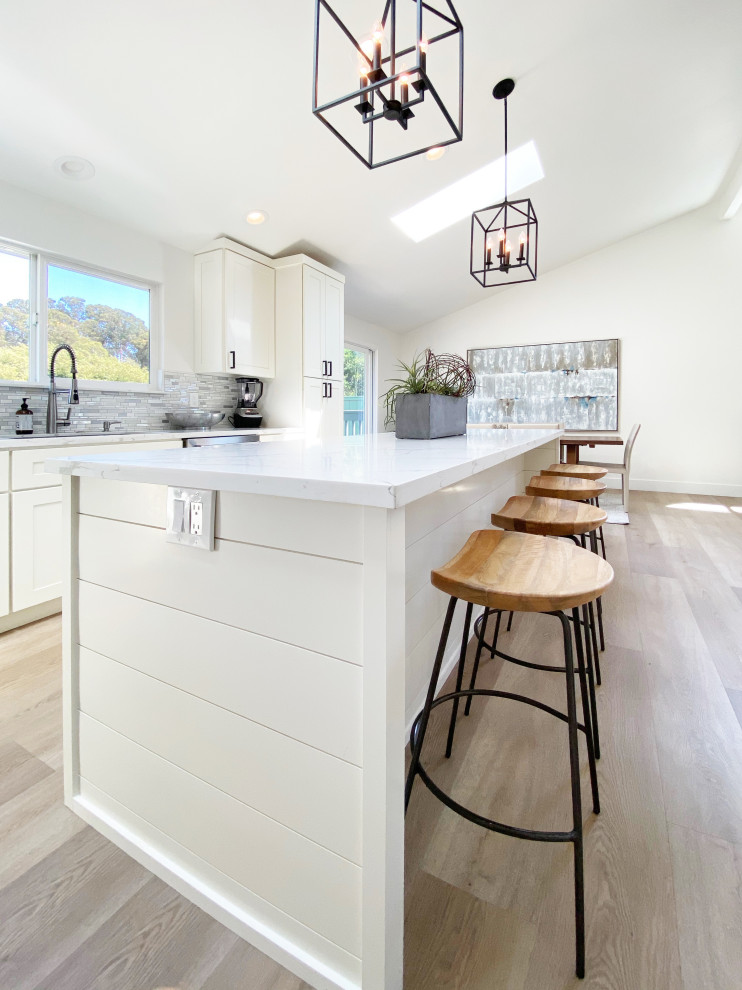 Example of a mid-sized transitional l-shaped vinyl floor, brown floor and vaulted ceiling eat-in kitchen design with an undermount sink, shaker cabinets, white cabinets, quartz countertops, multicolored backsplash, mosaic tile backsplash, stainless steel appliances, an island and white countertops