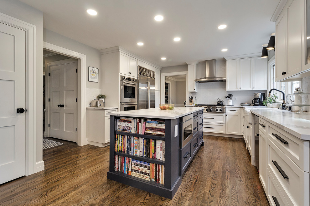 This is an example of a transitional l-shaped kitchen in Portland with a farmhouse sink, shaker cabinets, white cabinets, stainless steel appliances, dark hardwood floors, with island, brown floor and white benchtop.