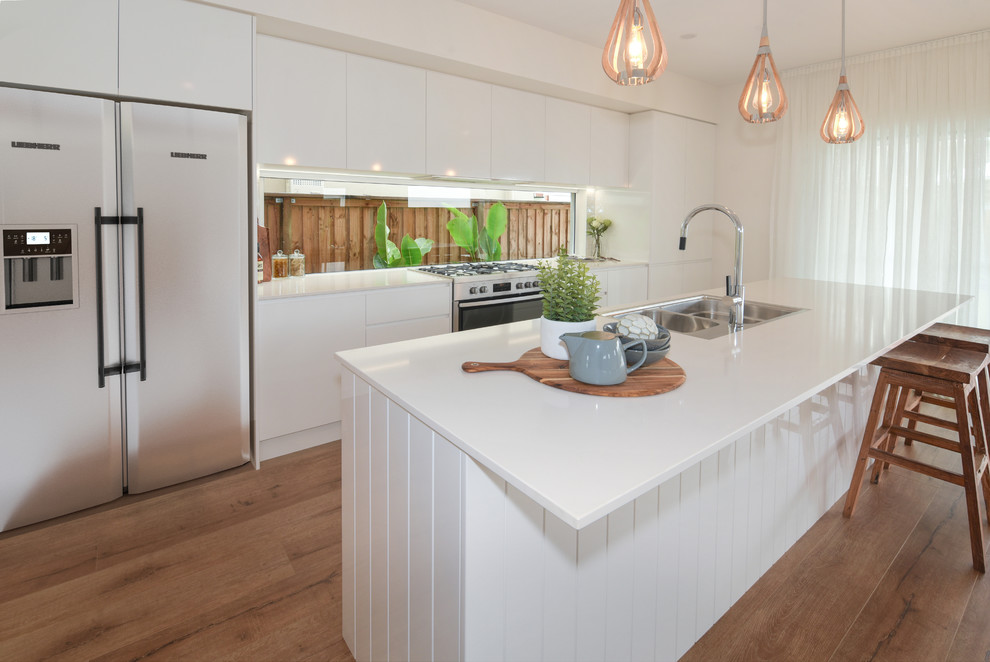 This is an example of a large beach style galley kitchen in Sunshine Coast with a drop-in sink, white cabinets, quartz benchtops, stainless steel appliances, with island, brown floor, white benchtop, flat-panel cabinets, window splashback and medium hardwood floors.