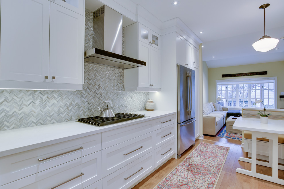This is an example of a mid-sized transitional galley kitchen in Toronto with an undermount sink, shaker cabinets, white cabinets, glass benchtops, grey splashback, stone tile splashback, stainless steel appliances, medium hardwood floors and no island.