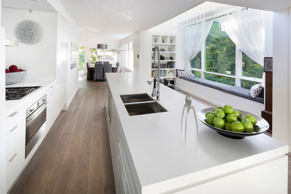 Mid-sized contemporary galley open plan kitchen in Sydney with flat-panel cabinets, stainless steel appliances, an undermount sink, white cabinets, quartz benchtops, white splashback, glass sheet splashback, light hardwood floors, brown floor and white benchtop.