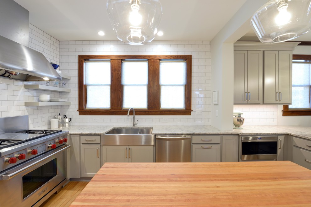 Mid-sized transitional light wood floor and brown floor enclosed kitchen photo with a farmhouse sink, shaker cabinets, gray cabinets, wood countertops, white backsplash, porcelain backsplash, stainless steel appliances, an island and brown countertops