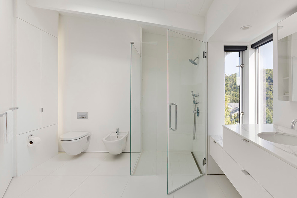 Photo of a mid-sized modern master bathroom in San Francisco with an undermount sink, a wall-mount toilet, flat-panel cabinets, white cabinets, marble benchtops, a drop-in tub, a curbless shower, white tile, porcelain tile, white walls, porcelain floors, white floor and white benchtops.
