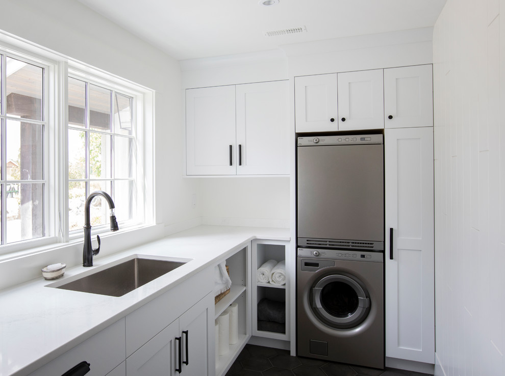 This is an example of a country galley dedicated laundry room in Other with shaker cabinets, white cabinets, marble benchtops, white walls, terra-cotta floors, a stacked washer and dryer, black floor and an undermount sink.
