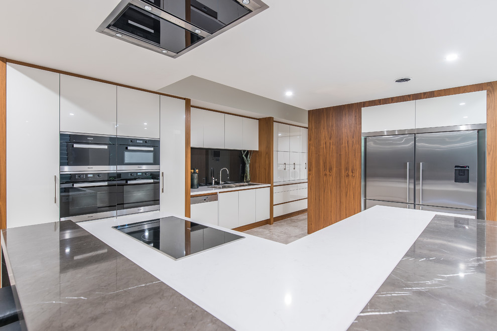 Contemporary u-shaped kitchen in Perth with a drop-in sink, flat-panel cabinets, white cabinets, black splashback, glass sheet splashback, stainless steel appliances, with island, grey floor and white benchtop.