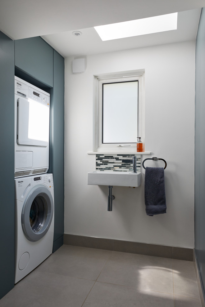 Design ideas for a mid-sized transitional utility room in Surrey with a single-bowl sink, flat-panel cabinets, blue cabinets, blue splashback, mosaic tile splashback, white walls, porcelain floors, a stacked washer and dryer, beige floor and recessed.