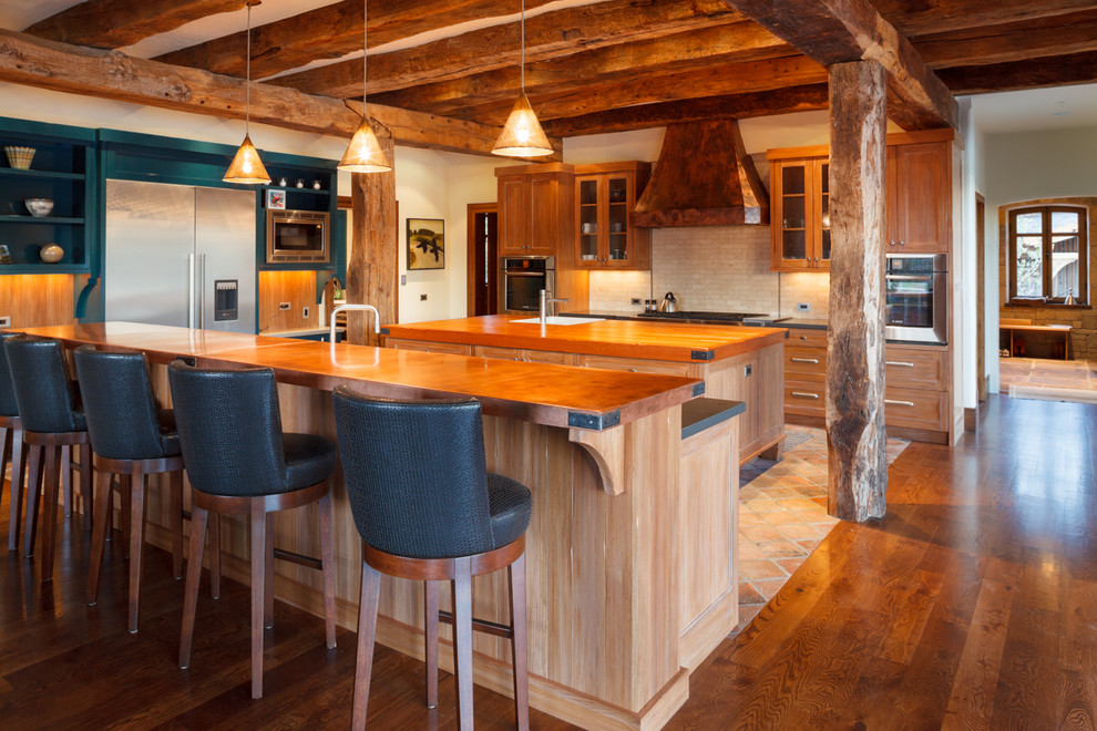Eat-in kitchen - large rustic l-shaped medium tone wood floor and brown floor eat-in kitchen idea with a farmhouse sink, shaker cabinets, medium tone wood cabinets, soapstone countertops, beige backsplash, stone tile backsplash, stainless steel appliances and two islands