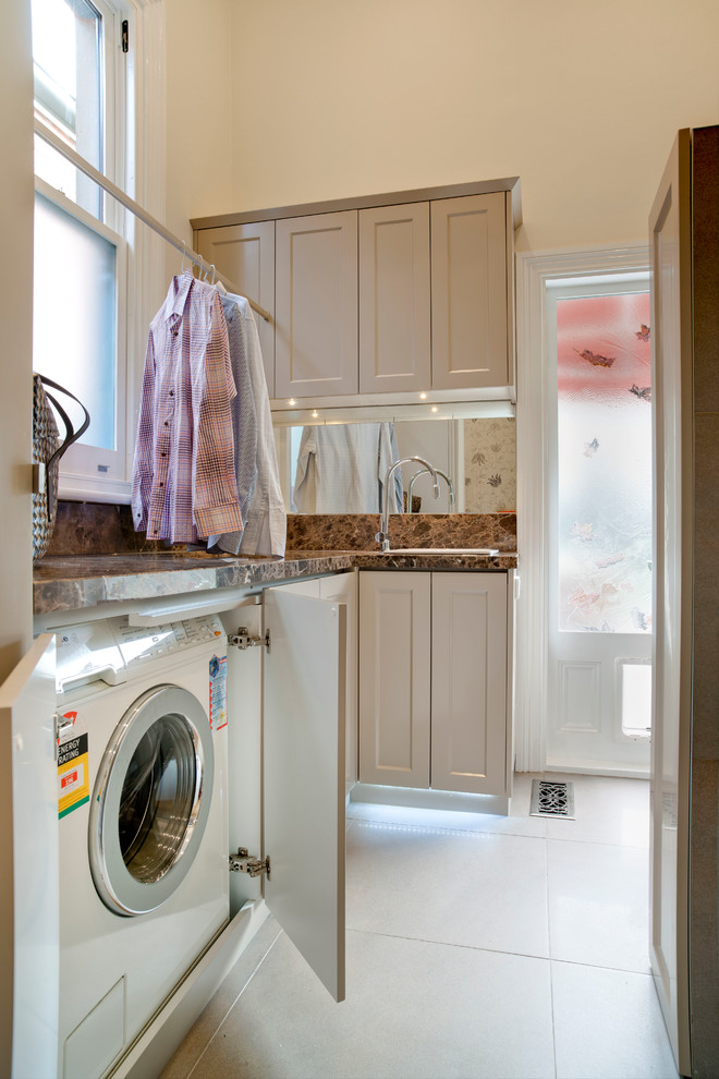 Photo of a small traditional u-shaped utility room in Melbourne with a drop-in sink, shaker cabinets, granite benchtops, white walls, porcelain floors, a concealed washer and dryer and beige cabinets.