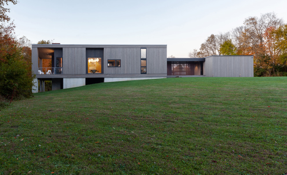 Foto della facciata di una casa piccola grigia moderna a un piano con rivestimento in legno, copertura in metallo o lamiera, tetto grigio, pannelli sovrapposti e terreno in pendenza