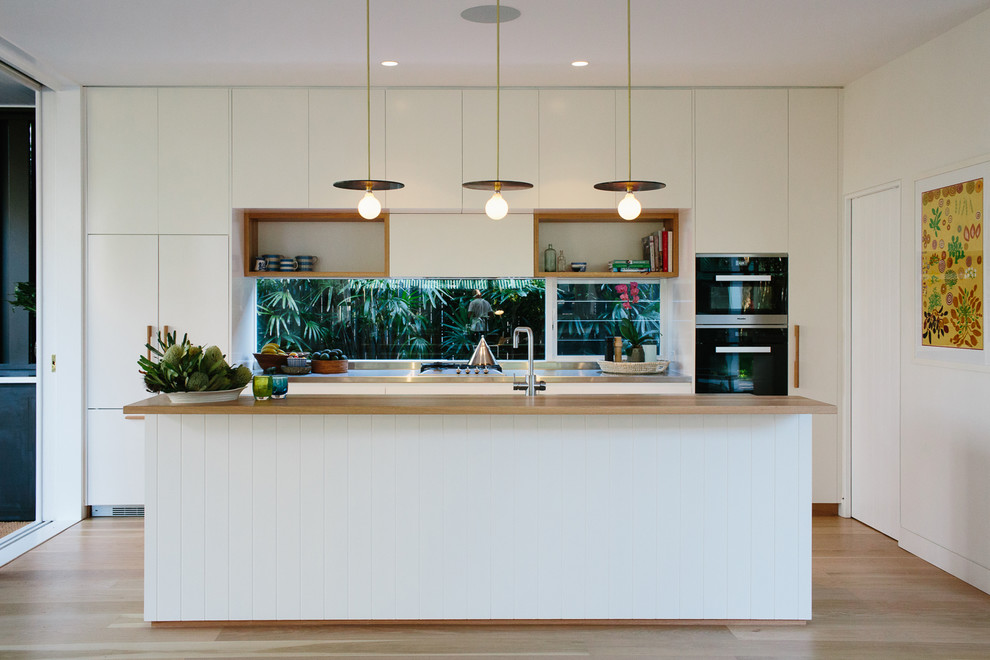 Photo of a mid-sized contemporary galley eat-in kitchen in Other with a double-bowl sink, recessed-panel cabinets, white cabinets, wood benchtops, white splashback, window splashback, black appliances, light hardwood floors, with island and white benchtop.